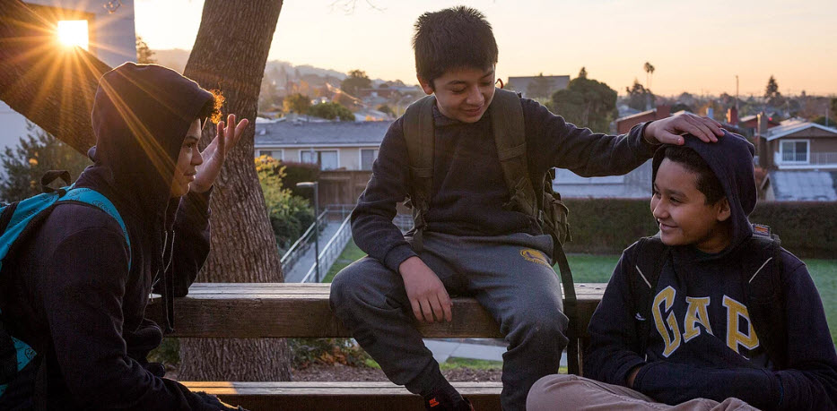 Three students interacting outside on a picnic table with sun setting in the background